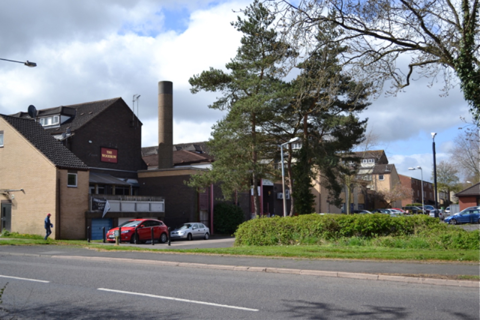 The Woodrow pub and Woodrow Centre behind tall pine trees, seen from Studley Road